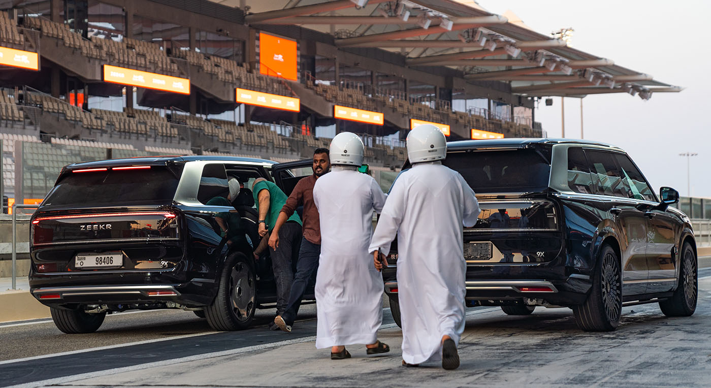Men in traditional white kandura attire next to two black luxury Zeekr 009 minivans at a race track pit lane.